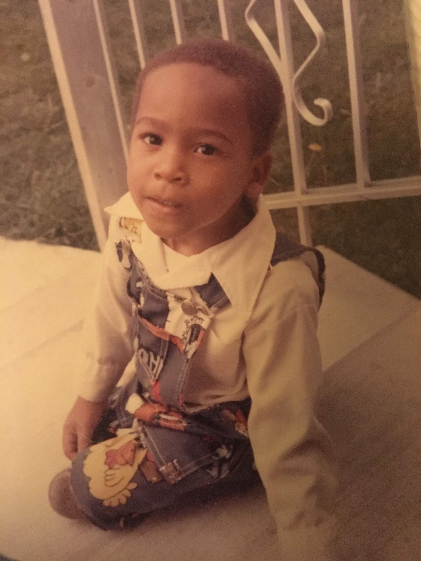 Eli Davis as a small boy in overalls and a white shirt, sitting on a porch behind white ironwork, looking at the camera