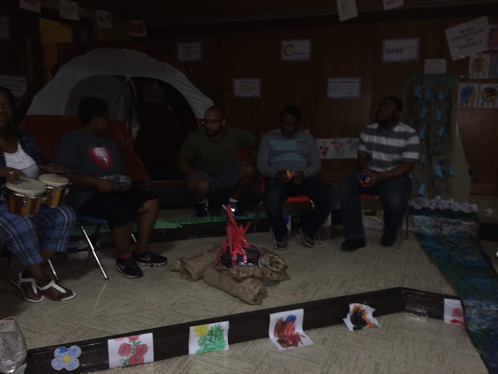 Students and staff gathered around the paper campfire inside the classroom campsite, drums in hand