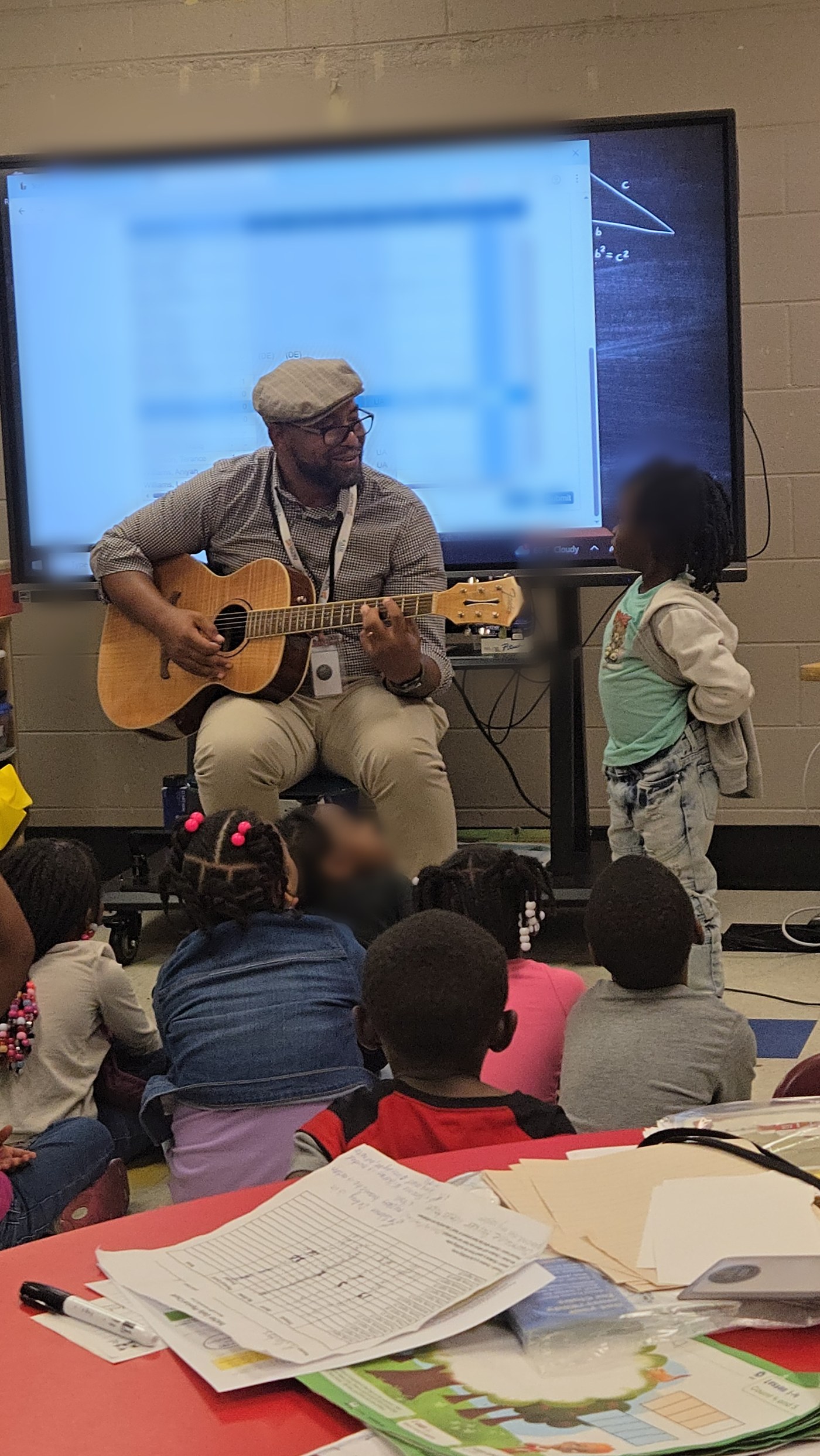 Eli Davis seated with an acoustic guitar playing for young students gathered on a classroom carpet; one student stands beside him; the screen behind him is blurred