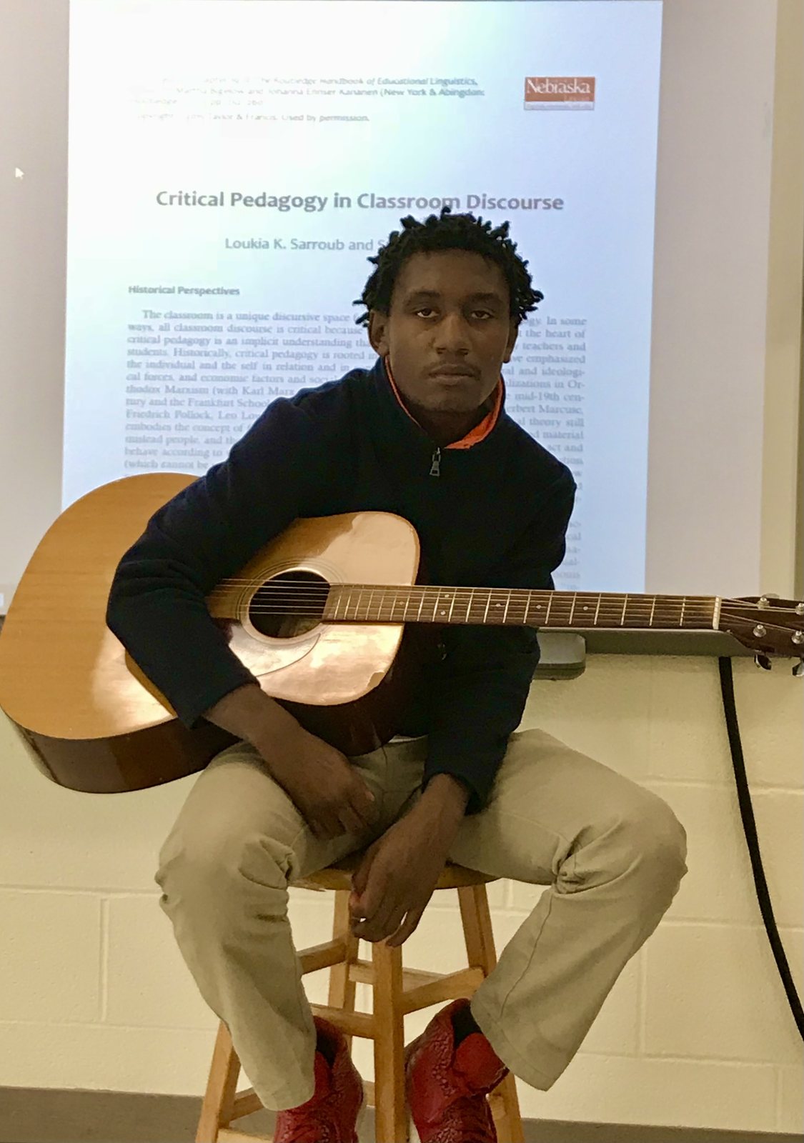 A student seated on a stool holding the acoustic guitar, a projected article on critical pedagogy behind him