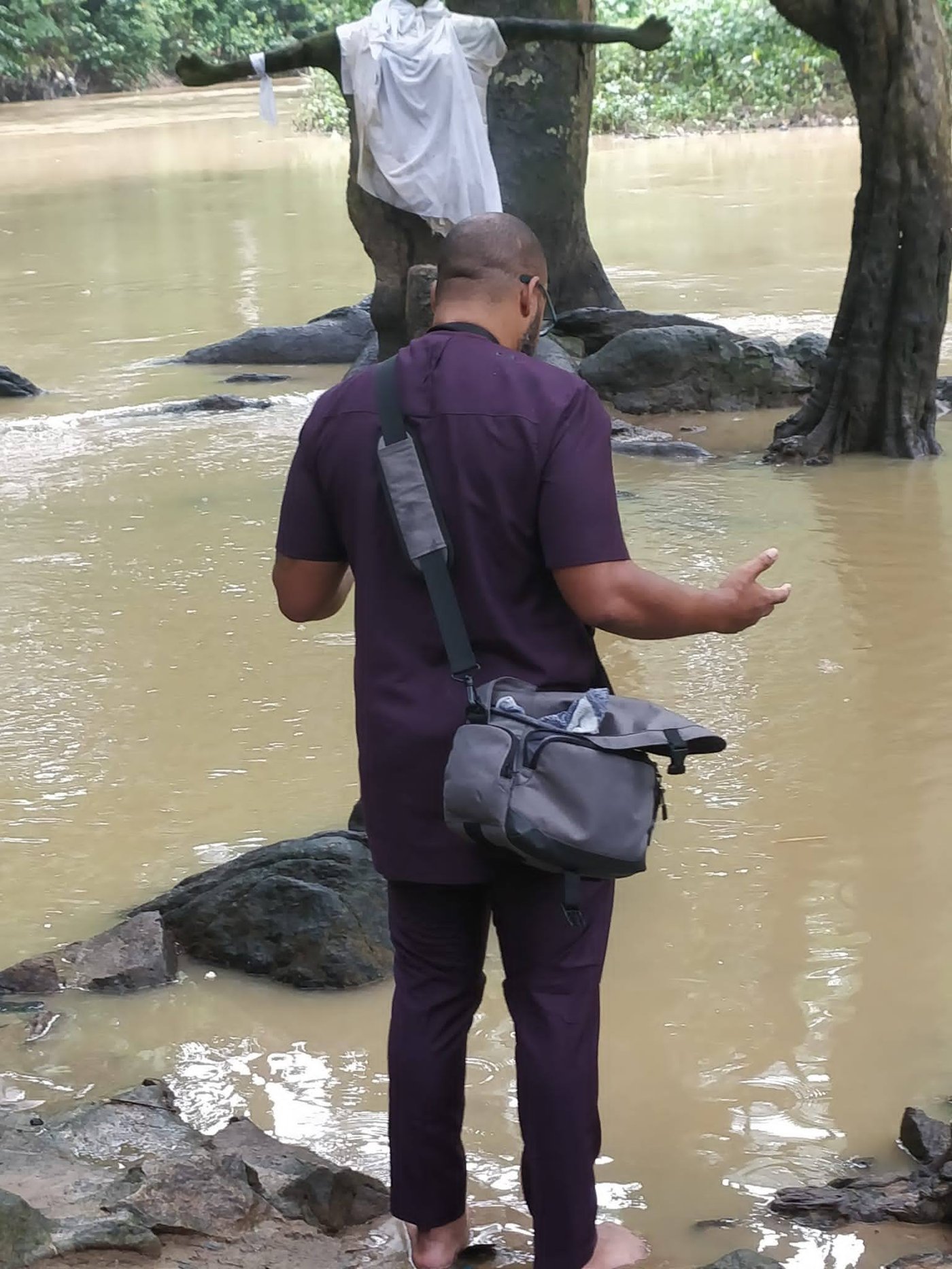 Eli Davis from behind, barefoot at the edge of the Osun River in Osogbo, Nigeria, hand open in prayer, a white-robed shrine figure across the water
