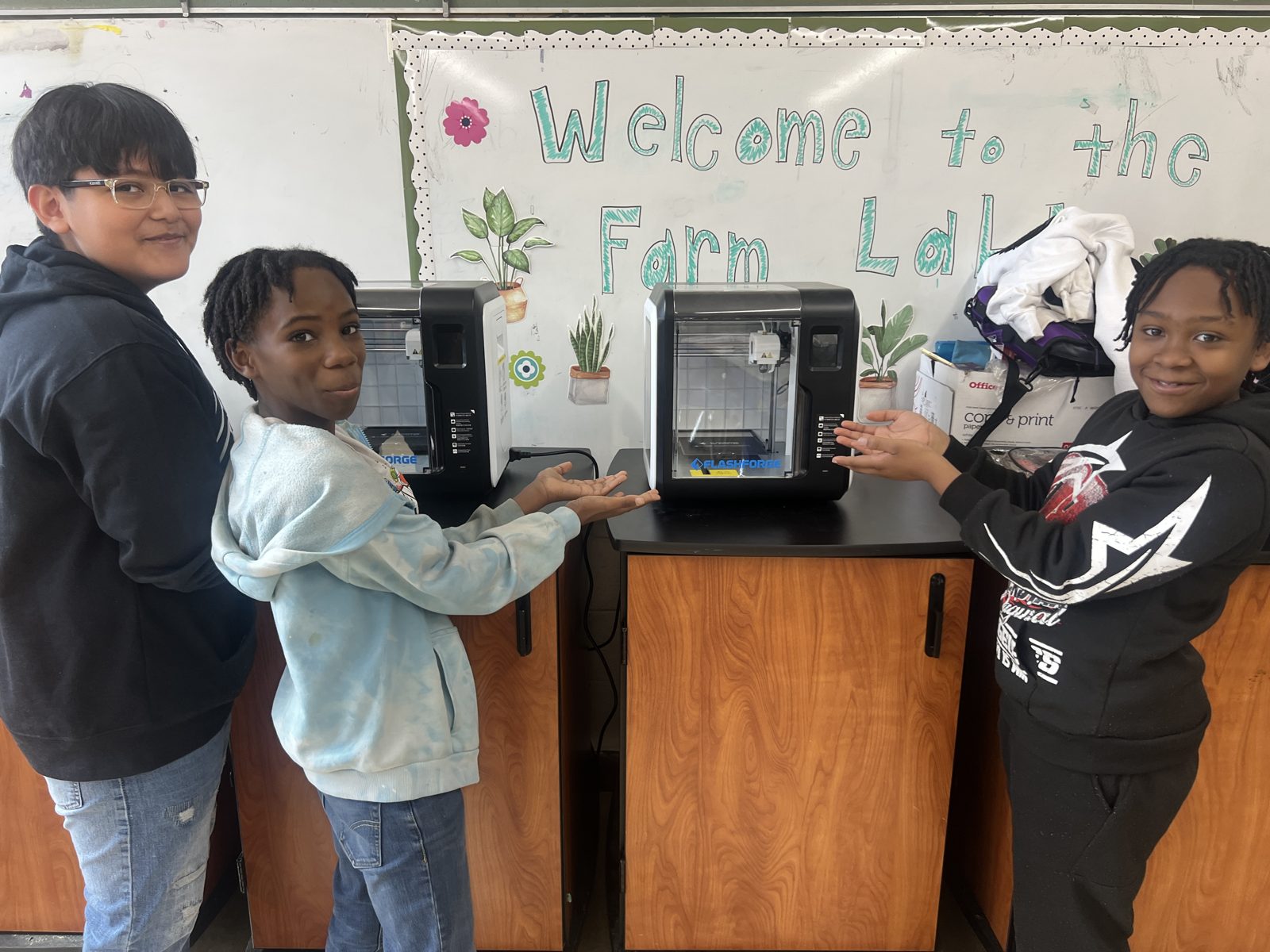 Three smiling students of the Cosmic STEM Club present two 3D printers